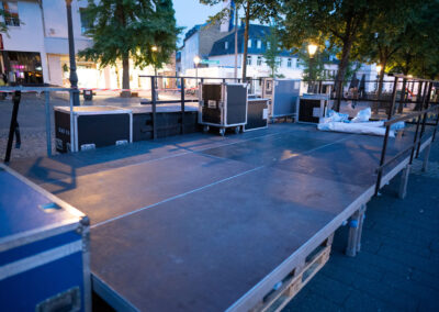 Eine Bühnenkonstruktion steht für den weiteren Aufbau bereit. Die Bühnenelemente und Absturzsicherungen sind angebracht, auf der Bühne stehen Flightcases mit Material.