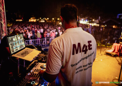 Ein DJ von M4E auf einem Open Air Bühne bei Nacht. Vor ihm ein Mischpult mit zwei CD-Playern und ein Laptop Computer zum abspielen von Musik für die tanzenden Menschen.