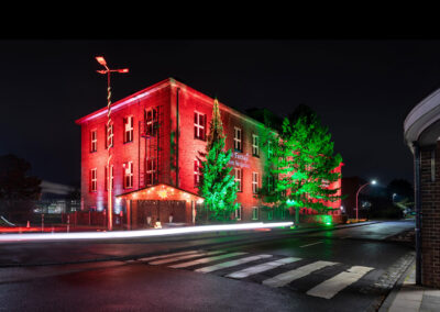 Eine rot angestrahlte zweistöckige Fassade. Die beiden Tannen davor sind in grünes Licht getaucht. Eine gelungene Architekturbeleuchtung.
