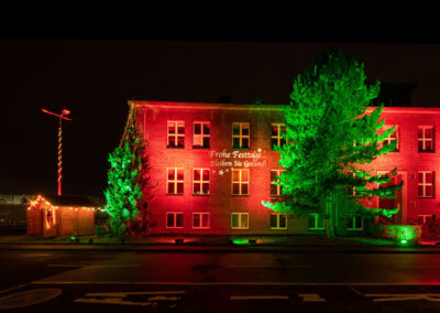 Architekturbeleuchtung. Eine rot angestrahlte zweistöckige Fassade. Die beiden Tannen davor sind in grünes Licht getaucht, was einen schönen Kontrast ergibt.