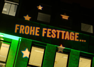 Gobo Projektion "Frohe Festtage" auf der Fassade des Baustoffhandel Henrich in Siegburg. Mit einem Gobo können Schrift und Ornamente an Fassaden projiziert werden.