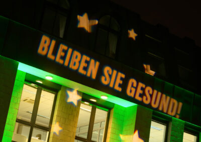 Gobo Projektion "Bleiben Sie gesund" auf der Fassade des Baustoffhandel Henrich in Siegburg. Mit einem Gobo können Schrift und Ornamente an Fassaden projiziert werden.