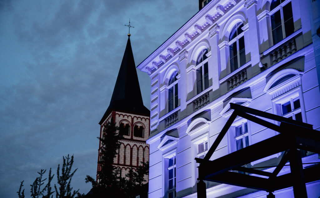 Violett illuminierte Gründerzeitfassade vor der Siegburger Kirche. Eine Installation von M4E Veranstaltungstechnik.